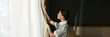 © pressmaster - Woman in housekeeping uniform opening curtains in well-lit room. She is looking out of window while revealing natural light into space we can see