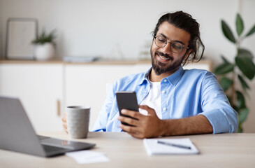 Poster - Smiling Indian Employee Using Smartphone And Having Coffee In Office, Enjoying Break At Work, Texting With Friends Or Browsing Social Networks, Sitting At Desk With Laptop, Selective Focus, Free Space