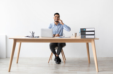 Poster - Corporate Communication. Smiling Arab Male Talking On Cellphone Working On Pc At Office, Positive Guy Sitting At Desk With Pc Having Phone Conversation, Enjoying Job, Free Copy Space, Full Body Length
