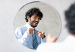 © Prostock-studio - You're handsome. Confident indian businessman pointing at mirror in bathroom with two fingers, man wearing shirt and smiling to his reflection, free space