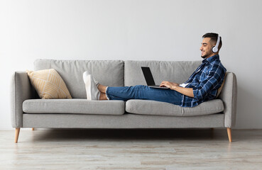 Aufkleber - Happy Arab guy resting sitting on couch at home, watching movie on laptop, using pc in wireless headset. Cheerful man playing video games typing on keyboard relaxing on weekend in living room