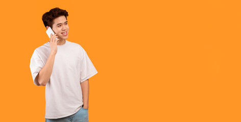 Poster - Always In Touch. Portrait of casual smiling asian guy talking on his smartphone, looking at copy space, standing and posing isolated over orange studio background, banner. Communication, Phone Call
