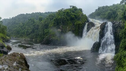 Poster - A powerful waterfall cascades through lush, dense rainforest, creating mist over rocky terrain with a wide river flowing below.