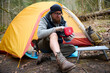 © Василь Івасюк - Hiker man with injured leg using first aid kit near the tent in the forest. A stressed trekker complaining alone in the mountain with broken knee.
