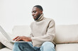 © SHOTPRIME STUDIO - Young African man working on a laptop at home, wearing a stylish beige sweater and relaxed jeans, exuding a focused and content emotion in a cozy environment