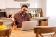 © Graphicroyalty - Smiling man working from home on a laptop, sitting at a wooden table. Cozy living room with warm lighting, bookshelves, and a bowl of fresh fruits, creating a relaxed yet productive atmosphere.