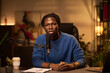 © pressmaster - Portrait of an African American man sitting at desk, speaking into microphone, using notes, engaging in conversation. Background includes blurred abstract artwork