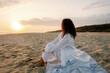 © Westend61 - Woman in white outfit sitting on a sandy beach at sunset, looking towards the horizon
