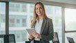 © Jari.art - Confident businesswoman in gray suit holds tablet, smiling in modern office setting with large windows