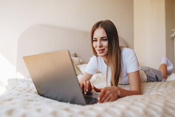 Poster - Young woman using laptop while lying on bed in cozy bedroom during the day