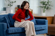 © Parichat - Asian woman experiencing severe abdominal pain while sitting on a sofa at home, potential health issue requiring medical attention