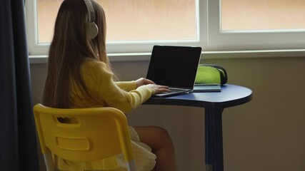 Aufkleber - A young girl is sitting at a desk, using a laptop and wearing headphones. She is in an indoor setting by a window, engaged in learning or entertainment on her computer. The scene is bright and focused
