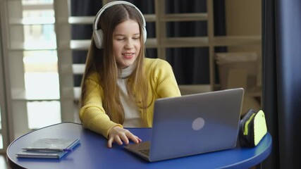 Aufkleber - A young girl sits at a round blue table, wearing headphones and using a laptop. She is smiling and appears to be engaged in a video call. Notepads and a pencil case are on the table.