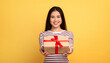 © Prostock-studio - A cheerful Asian young woman stands against a bright yellow background, holding a beautifully wrapped gift box with a red ribbon, ready to share her joy with someone special.