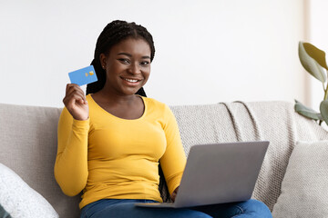 Poster - Easy Payment. Smiling Black Lady With Laptop And Credit Card At Home Making Online Payments Or Shopping In Internet, African Woman Sitting On Couch In Living Room And Looking At Camera, Free Space