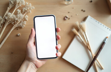 Poster - Hand holding modern smartphone, white screen. App template mockup. Workspace with dry plants, brushes, notebook, pen. Creative designer workspace. Social media, blog content, mobile app design. Blank