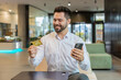 © Andrii Iemelianenko - Happy Caucasian businessman using bank credit card and smartphone for online shopping payments in modern office lobby. Business transaction. Smiling guy purchase with cellphone E-banking app service.
