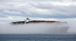 © MariaMedeiros - A container ship shrouded in a thick fog, sailing across the ocean under a cloudy sky.