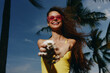 © SHOTPRIME STUDIO - Joyful woman smiling and playing with sand on a beach, wearing pink sunglasses and a yellow swimsuit against a blue sky with palm trees