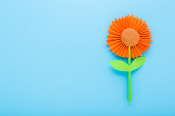 Wall Mural - Orange flower with green leaves created from colorful application paper on light blue table background. Pastel color. Closeup. Empty place for text. Top down view.