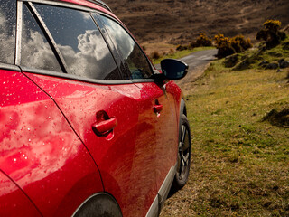 Wall Mural - A red family car is parked on a road by a field. The car is parked on a dirt road with a few trees in the background. The sky is cloudy. Travel and rural area tourism