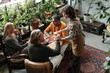© pressmaster - Group sitting around wooden table in cozy cafe surrounded by greenery, engaging in lively conversation and sharing tea while server hands small teacup to person wearing green sweater