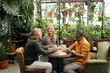 © pressmaster - Three people enjoying coffee at a wooden table in a lush green setting with many plants. Natural light filtering through large windows highlighting the cozy atmosphere