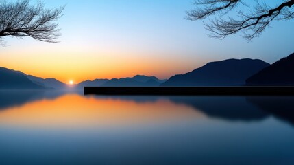 Wall Mural - The sun sets slowly beyond distant mountains, casting warm colors across a calm lake. Bare tree branches frame the view, enhancing the peaceful twilight atmosphere.