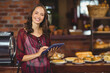 © WavebreakMediaMicro - Woman standing behind counter in bakery holding tablet and showing pastries beside coffee machine