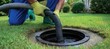 © dreamer - A worker uses a hose to clean or pump a residential septic tank in a well-maintained grassy yard.