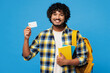 © ViDi Studio - Young smiling happy fun Indian boy student wears yellow casual clothes backpack bag hold books credit bank card look camera isolated on plain blue background. High school university college concept.