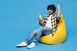 © ViDi Studio - Full body young Indian man he wear yellow checkered shirt casual clothes sit in bag chair hold in hand use mobile cell phone do winner gesture isolated on plain blue cyan background Lifestyle concept