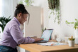 © Studio Marmellata - Young woman works from home on her laptop at a desk. She is smiling while typing, with headphones and plants around her. Remote work and online learning.