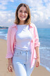 © Raquel - Smiling young woman walks barefoot along a sandy beach on a sunny day. She wears light clothes and enjoys the peaceful coastal atmosphere with waves, blue sky, and natural beauty around her. Vertical