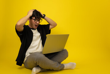 Young freckled man wearing glasses sitting with a surprised expression in front of his laptop on a yellow background with copy space