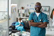 © pressmaster - African American doctor standing with arms crossed, smiling confidently in modern medical office, while a nurse sits on couch in background, busy with paperwork