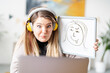 © Studio Marmellata - A woman with headphones is video conferencing, holding up a whiteboard with a silly self-portrait. She's making a funny face to match the drawing, trying to make her audience laugh..