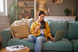 © Stockphotodirectors - A woman dressed in a yellow shirt sits comfortably on a couch, sipping from a mug while focused on her laptop. The room has natural light and a relaxed atmosphere.