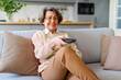 © Home-stock - Happy smiling senior woman sitting on couch in the living room, pointing tv remote controller to switch channels, watching favorite movie online series