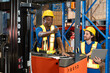 © yuttapon - Black male warehouse worker standing at forklift, pointing forward while explaining procedure to bearded colleague holding laptop. Both wearing helmet and vest inside organized storage facility.