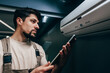 © fotofabrika - Technician inspects air conditioning unit while checking readings on a tablet in a modern indoor space