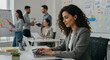 © Fauzan - Beautiful Middle Eastern Manager Sitting at a Desk in Creative Office. Young Stylish Female with Curly Hair Using Laptop Computer in Marketing Agency. Colleagues Working in the Background
