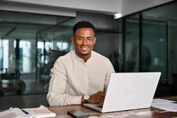 Portrait of smiling male company employee or entrepreneur in office. Happy young professional African American business man in glasses looking at camera sitting at work desk using computer.
