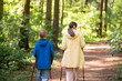 © kazakova0684 - Rear view of mother and 11-year-old son hiking with Nordic trekking poles through a sunny forest trail