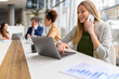 © Moon Safari - Businesswoman working on laptop and speaking on smart phone with professional colleagues collaborating in the background