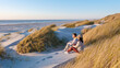 © Fokke Baarssen - Two people sit together on the soft dunes, surrounded by tall grass, gazing at the calm waves of the North Sea during golden hour in Blokhus Strand,Denmark