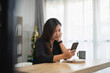 © WMSTUDIO - Young woman sitting at a wooden table in a bright room, using smartphone while relaxing, festive decorations in the background enhance the cozy atmosphere.
