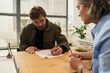 © Mediaphotos - Middle aged Caucasian man sitting at desk signing paperwork with middle aged Caucasian female social worker in office environment, engaging in social services consultation
