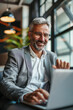 © Starmarpro - Smiling mature businessman with silver hair works on a laptop in a modern office setting. He looks professional and approachable.