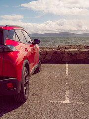 Wall Mural - A red car is parked in a parking lot next to a wall. The car is facing the ocean, and the sky is cloudy. Car park with stunning view on Galway bay and Burren mountains. Travel and tourism.